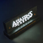 Lampe avec le logo du jeu vidéo Assassin's Creed, de la marque Neamedia Icons, allumée, en situation sur un bureau